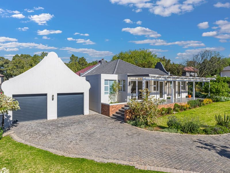 A well-maintained single-story home featuring a double garage with Cape Dutch style gables, a paved driveway, and a covered patio overlooking a landscaped garden.