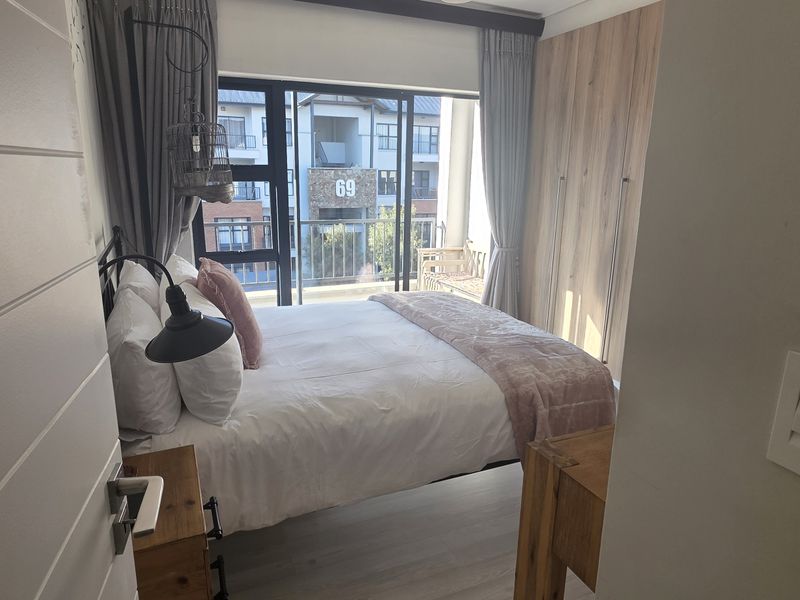 A well-lit bedroom featuring modern built-in cupboards, laminate flooring, and a sliding glass door leading to a balcony.