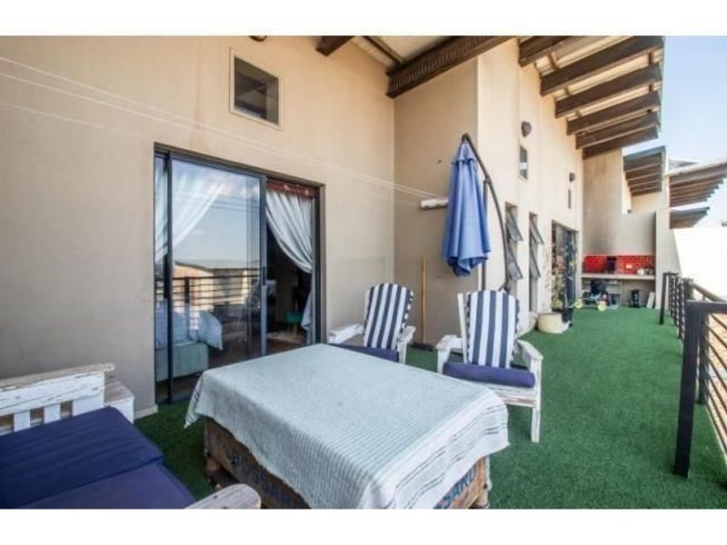 Spacious covered balcony featuring artificial grass, exposed timber rafters, sliding door access, and a built-in outdoor braai area.