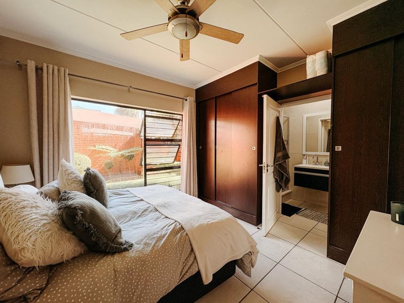 A well-maintained bedroom featuring built-in dark wood cupboards, tiled flooring, a ceiling fan, and an adjoining en-suite bathroom.