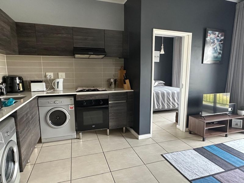 Modern open-plan kitchen featuring dark wood cabinetry, integrated appliances, and tiled flooring, seamlessly connecting to the adjacent bedroom area.