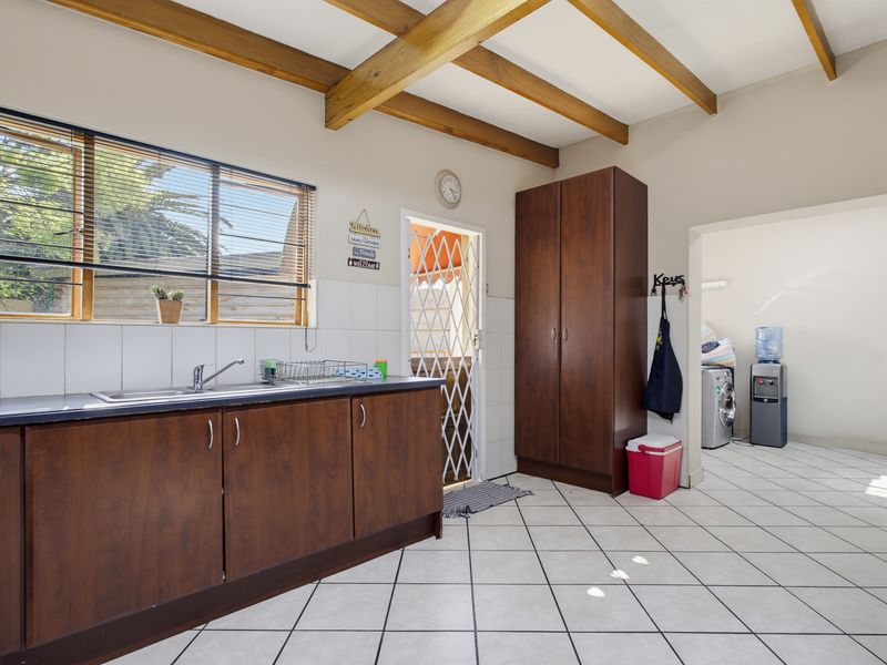 A functional scullery area featuring dark wood cabinetry, a stainless steel sink, tiled splashback, and exposed timber ceiling beams, with an adjacent utility space.