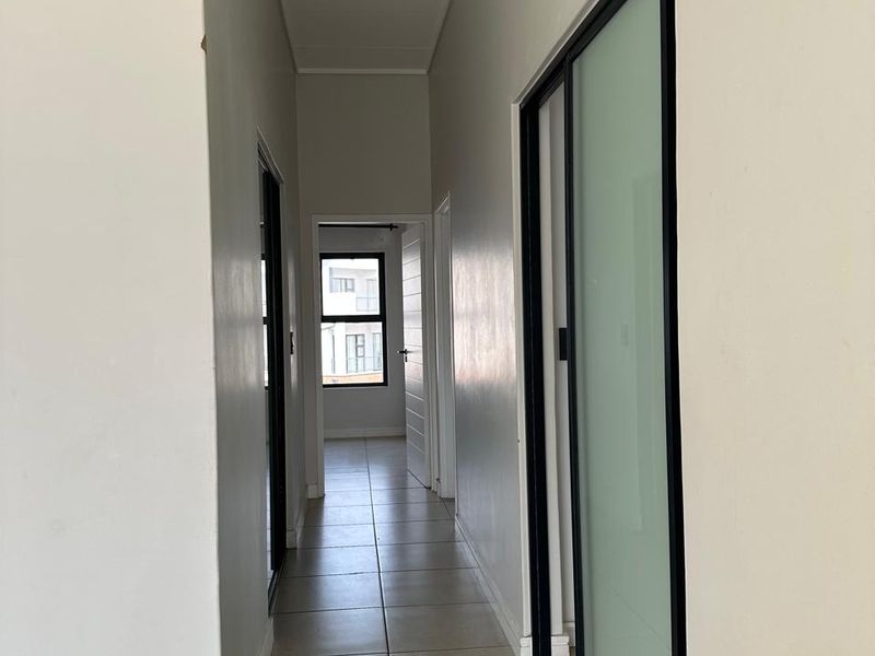 A clean, modern hallway featuring neutral tiled flooring, minimalist black-framed sliding doors, and crisp white walls leading to a bright bedroom area.