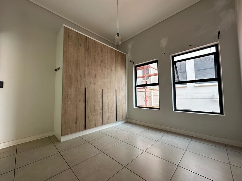 A bright, empty bedroom featuring modern wood-finish built-in wardrobes, neutral tiled flooring, and black aluminium windows. The walls require final painting and finishing touches.