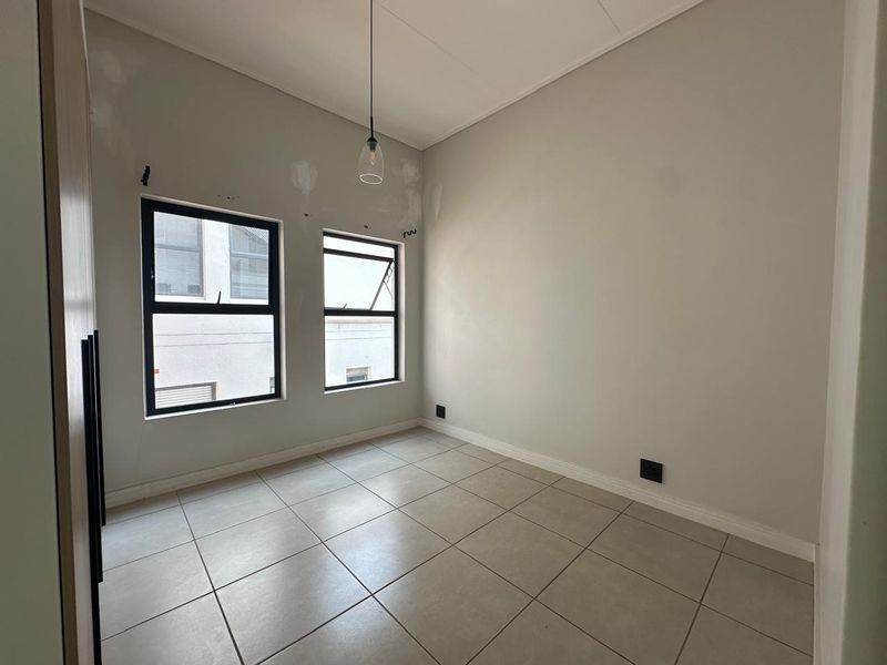 An empty bedroom featuring tiled flooring, built-in cupboards, and modern aluminium windows, currently requiring minor wall finishing and paint touch-ups.