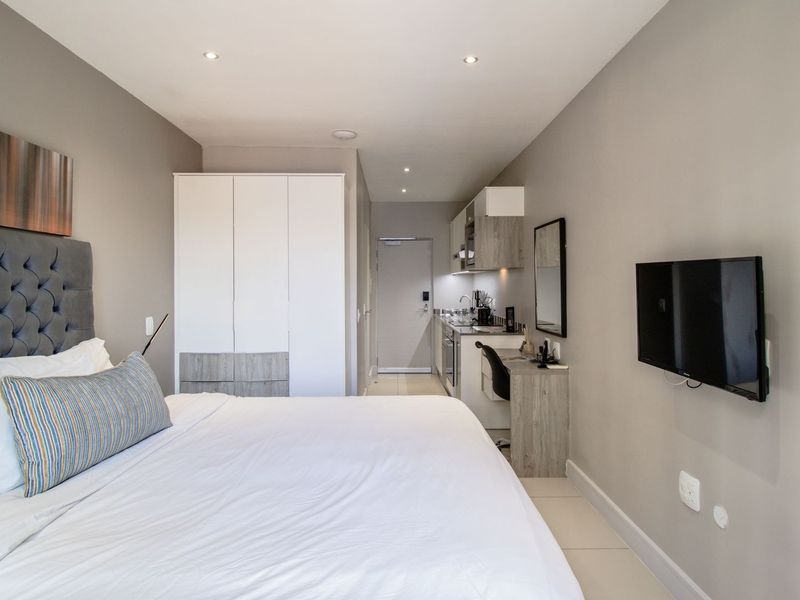 A modern studio bedroom featuring a built-in wardrobe, kitchenette with cabinetry, and tiled flooring, offering a compact and functional living space.