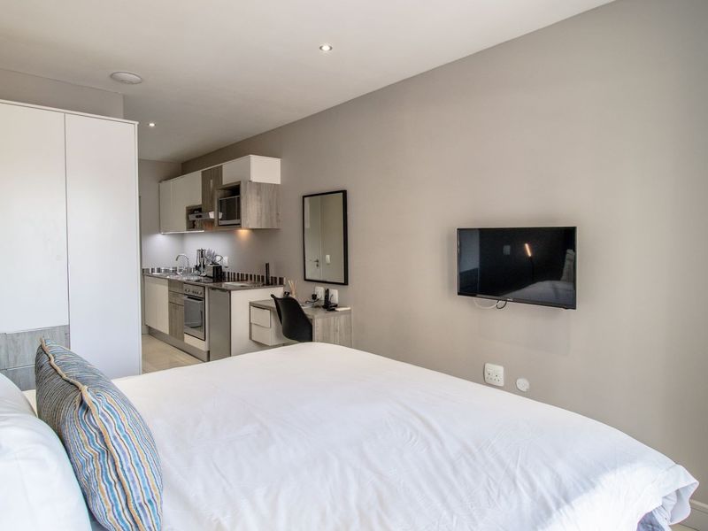 A modern open-plan studio apartment featuring a compact kitchenette, built-in wardrobe, integrated desk space, and a wall-mounted television.