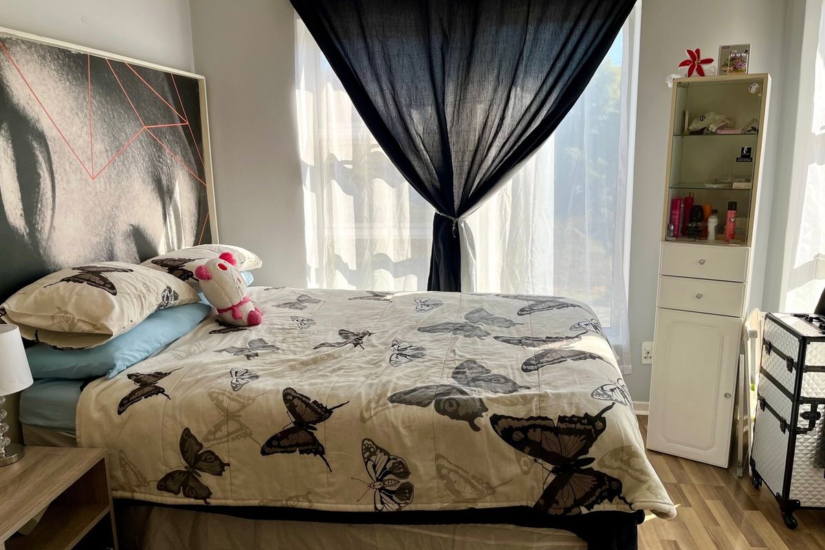A bedroom featuring laminate flooring and a built-in storage unit, illuminated by natural light from a window with draped curtains.