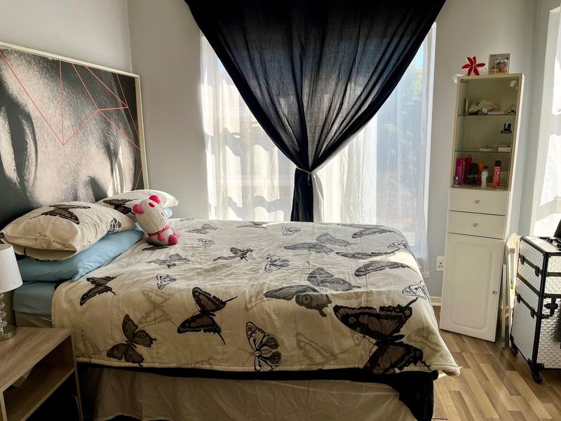 A bedroom featuring laminate flooring and a built-in storage unit, illuminated by natural light from a window with draped curtains.