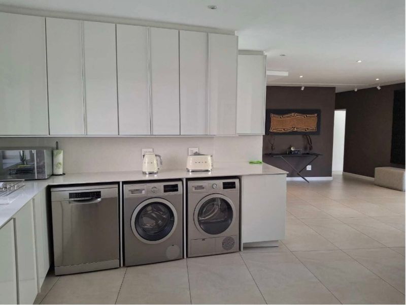 Modern kitchen featuring sleek white cabinetry, stone countertops, and integrated laundry appliances, seamlessly flowing into an open-plan living area with tiled floors.