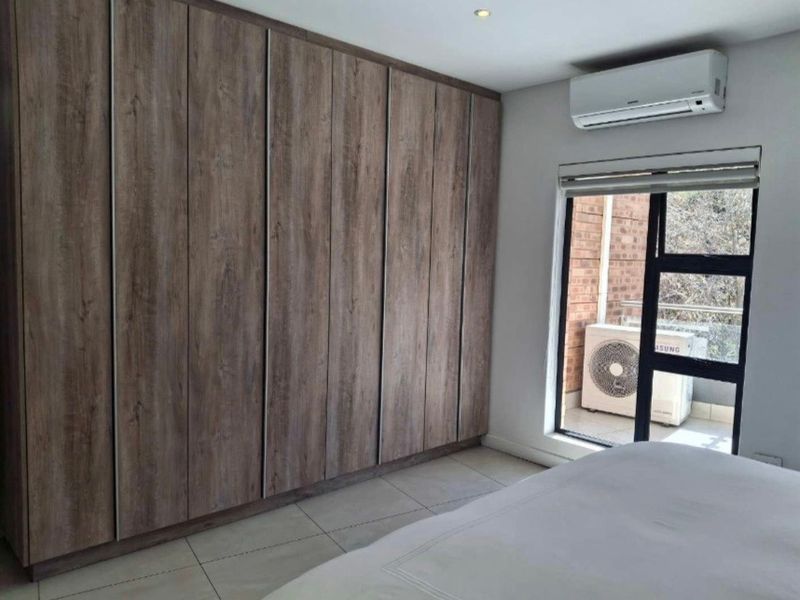 A well-maintained bedroom featuring extensive built-in wooden cupboards, tiled flooring, and a wall-mounted air conditioning unit with access to an outdoor balcony.