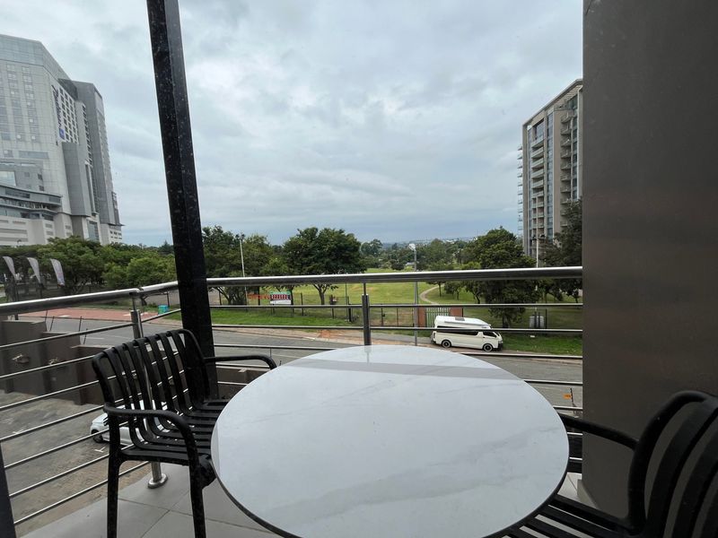 A covered balcony featuring a stainless steel balustrade and tiled flooring, offering an elevated view of the surrounding urban landscape and park area.