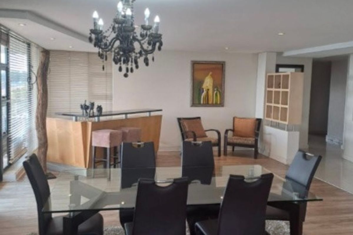 An open-plan living and dining area featuring a built-in bar counter, decorative chandelier, and large windows with venetian blinds.