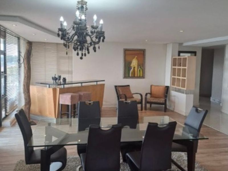 An open-plan living and dining area featuring a built-in bar counter, decorative chandelier, and large windows with venetian blinds.