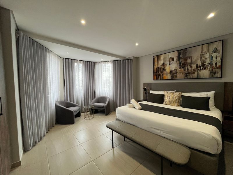 A spacious, modern bedroom featuring neutral tiled flooring, recessed ceiling lighting, and floor-to-ceiling curtains covering a curved window wall.