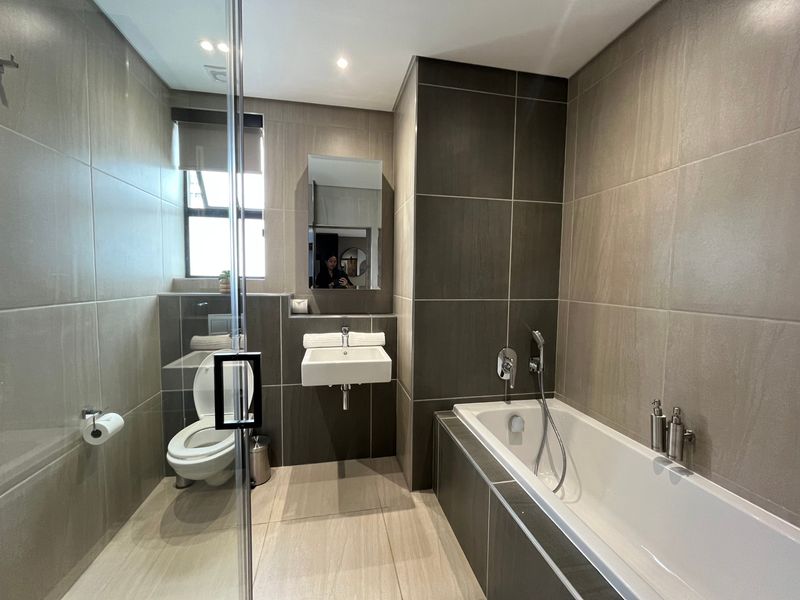 Modern bathroom featuring a built-in bathtub, wall-mounted basin, glass shower screen, and neutral floor-to-ceiling tiling with recessed lighting.