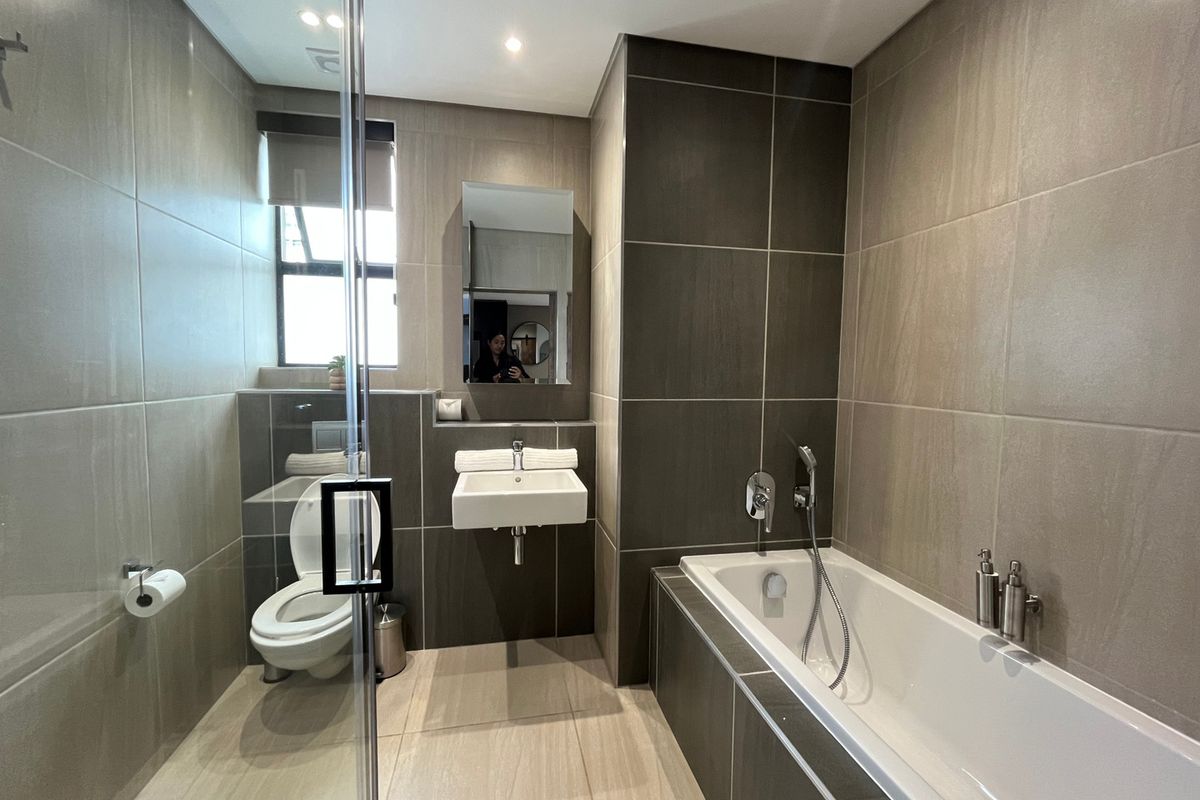 Modern bathroom featuring a built-in bathtub, wall-mounted basin, glass shower screen, and neutral floor-to-ceiling tiling with recessed lighting.