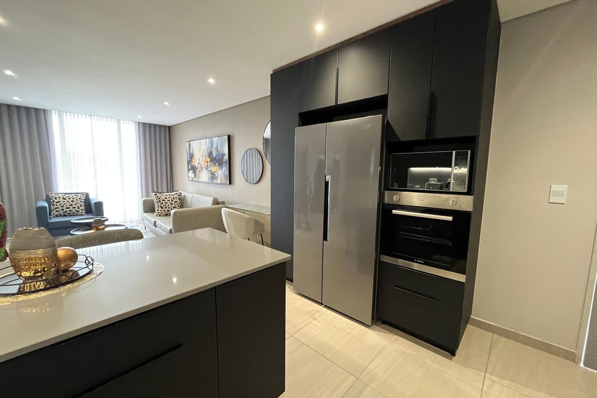 Modern open-plan living area featuring a sleek kitchen with dark cabinetry, stone countertops, integrated appliances, and recessed lighting, flowing into a bright lounge space.