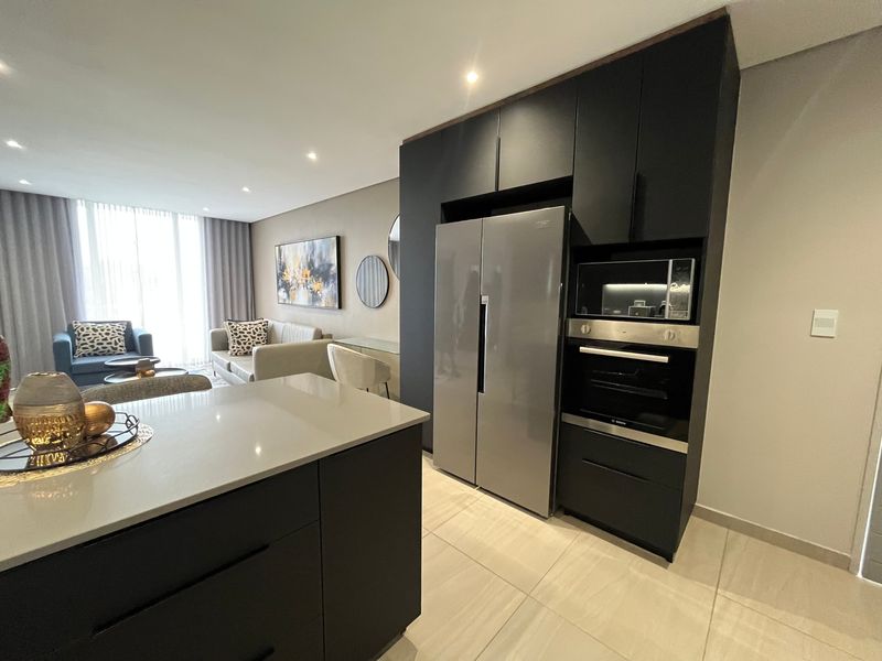 Modern open-plan living area featuring a sleek kitchen with dark cabinetry, stone countertops, integrated appliances, and recessed lighting, flowing into a bright lounge space.