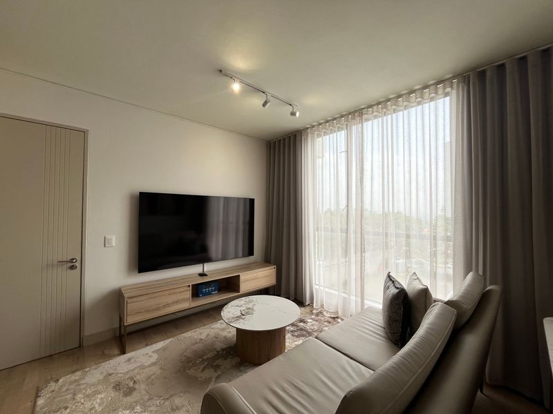 A modern, well-lit living room featuring track lighting, neutral wall finishes, and large windows with floor-to-ceiling sheer curtains.
