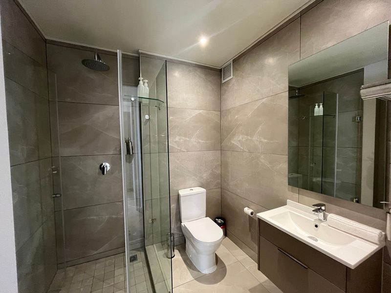Modern bathroom featuring large-format neutral wall tiles, a glass-enclosed walk-in shower with rain head, wall-mounted toilet, and a sleek vanity with integrated basin.