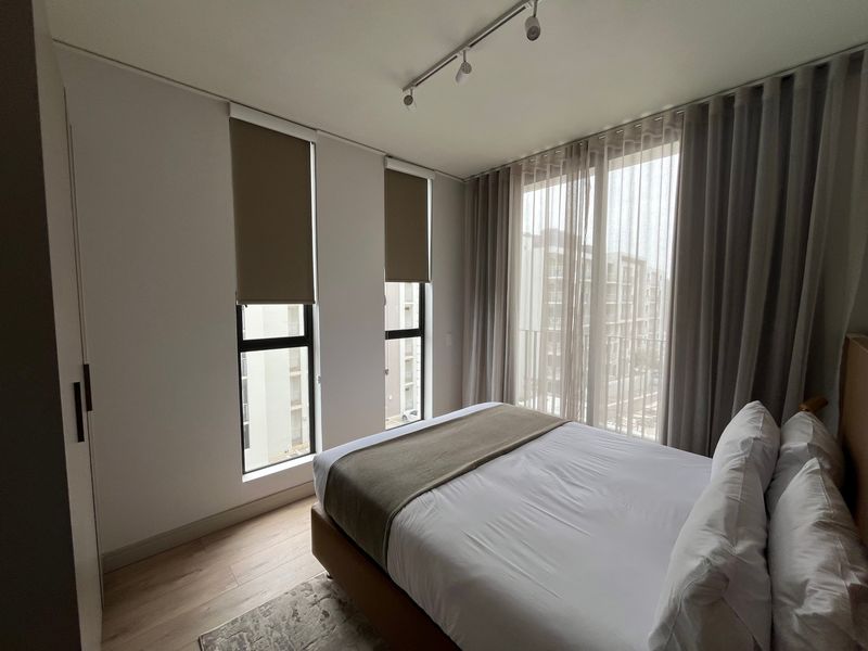 Modern bedroom featuring light laminate flooring, built-in wardrobe, track lighting, and large windows with roller blinds and sheer curtains.