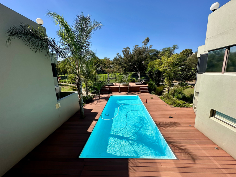 A well-maintained outdoor pool area featuring a rectangular swimming pool surrounded by timber decking and landscaped garden views.