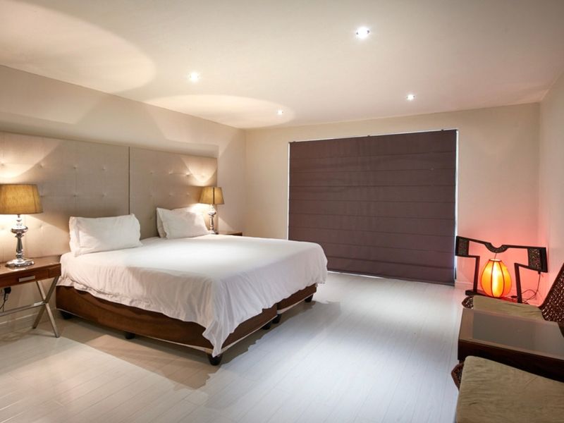 A spacious bedroom featuring recessed downlighting, a large upholstered headboard, and a dark roman blind covering the window.