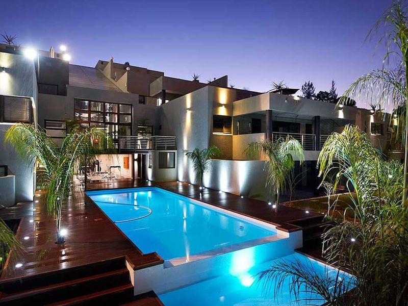 A beautifully illuminated swimming pool area featuring modern timber decking, integrated lighting, and a multi-level architectural facade with balconies.