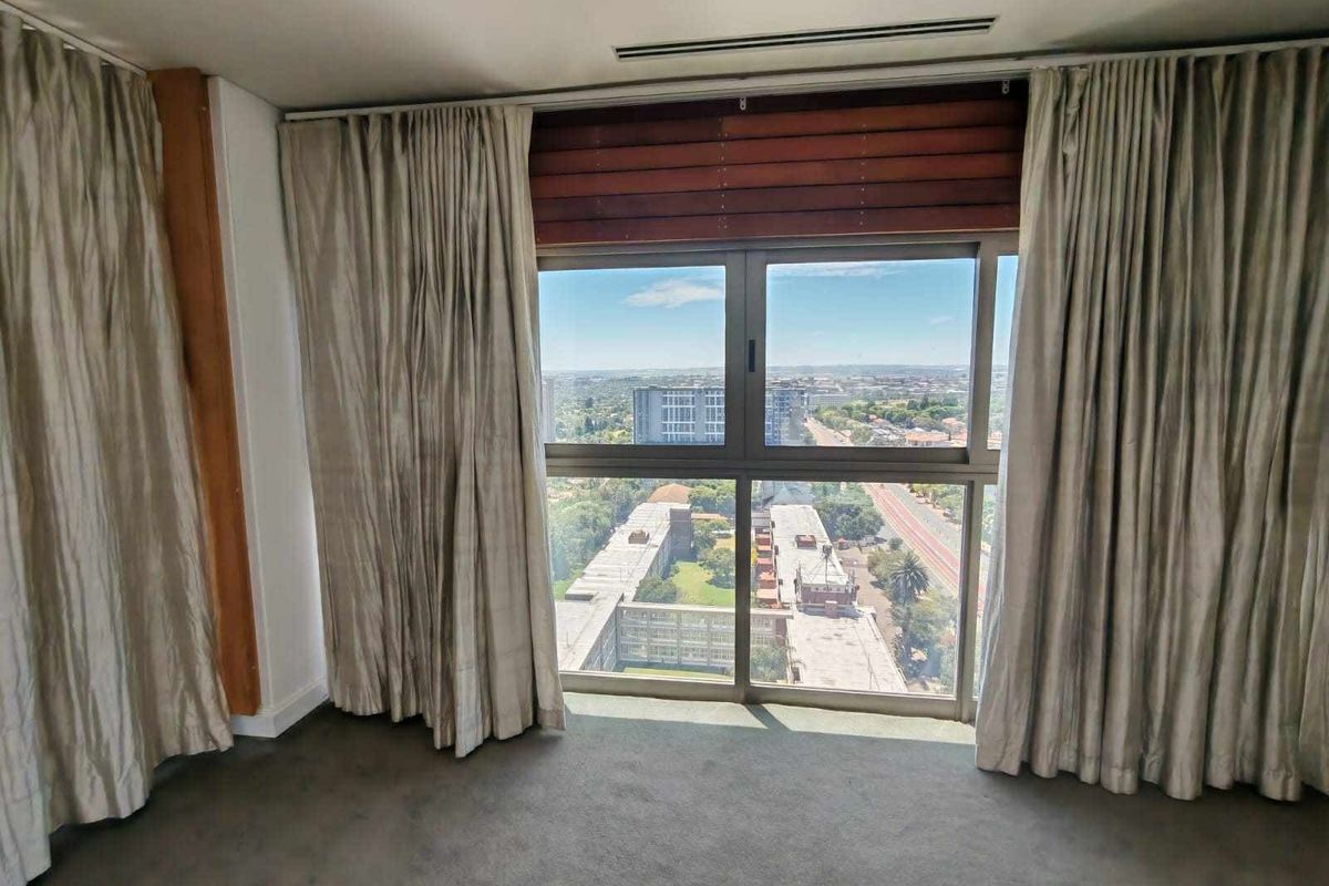 A spacious, empty bedroom featuring large windows with expansive city views, neutral carpeting, and recessed ceiling lighting.