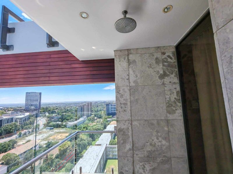 A modern balcony featuring a glass balustrade, stone wall cladding, and a ceiling-mounted shower head, offering expansive urban views.