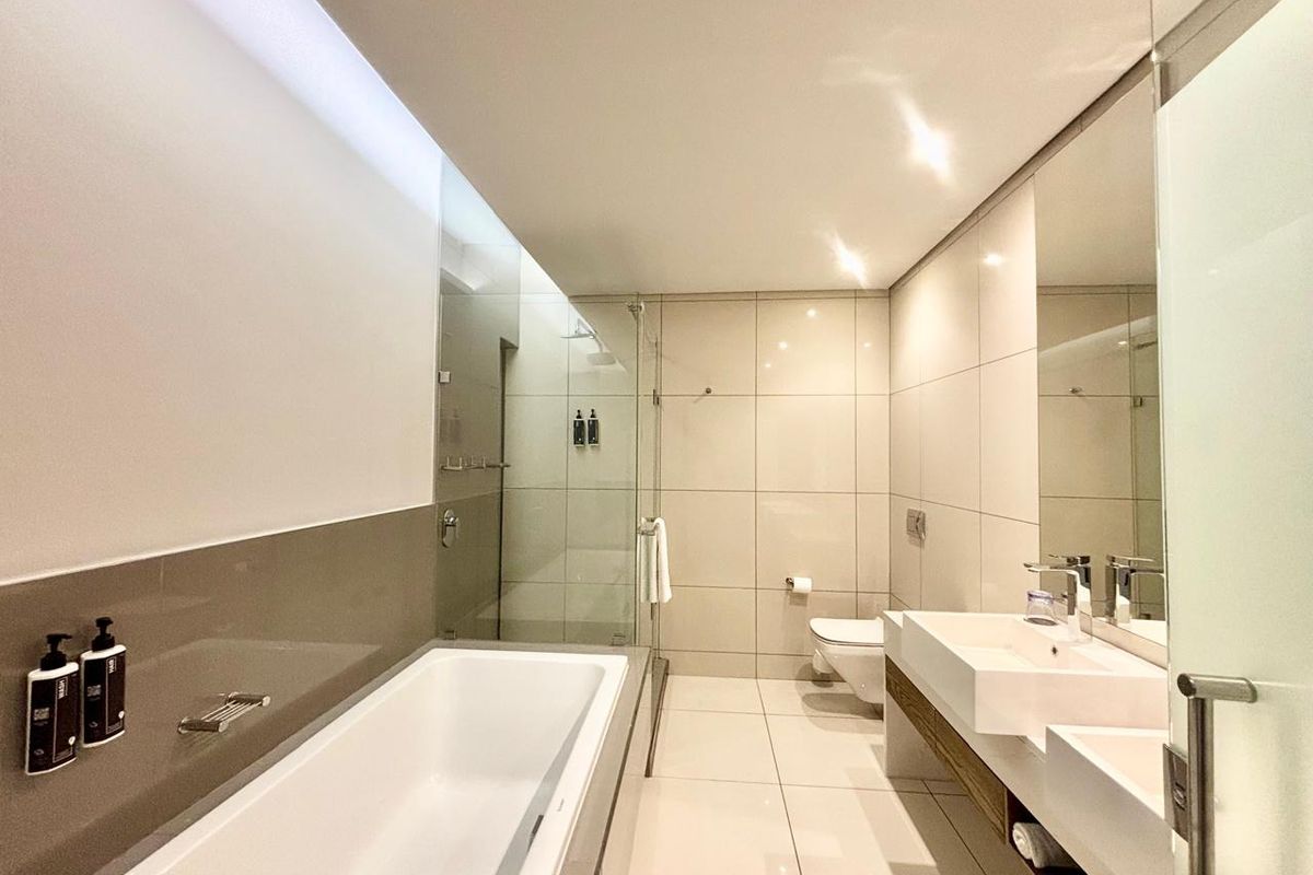 Modern hotel bathroom featuring a built-in bathtub, glass-enclosed shower, wall-mounted toilet, and a sleek double vanity with large mirror.