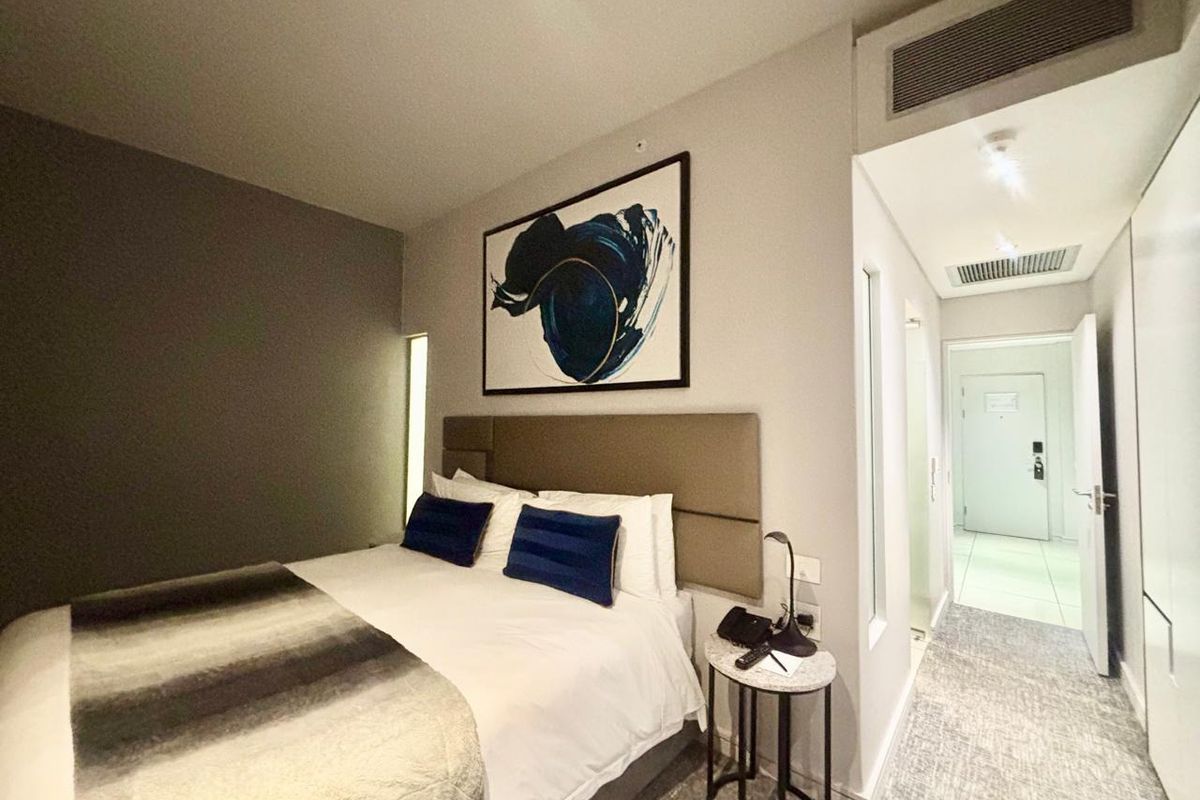 A modern hotel bedroom featuring a neutral color palette, upholstered headboard, and integrated ducted air conditioning system.