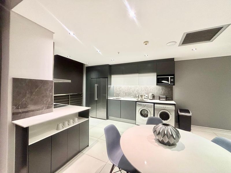 Modern open-plan kitchen featuring sleek dark cabinetry, stone backsplash, integrated appliances, and recessed lighting, seamlessly flowing into the dining area.