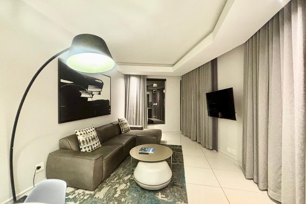 A modern hotel living area featuring tiled floors, recessed ceiling lighting, and a wall-mounted television with floor-to-ceiling drapery.