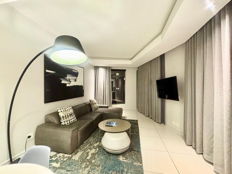 A modern hotel living area featuring tiled floors, recessed ceiling lighting, and a wall-mounted television with floor-to-ceiling drapery.