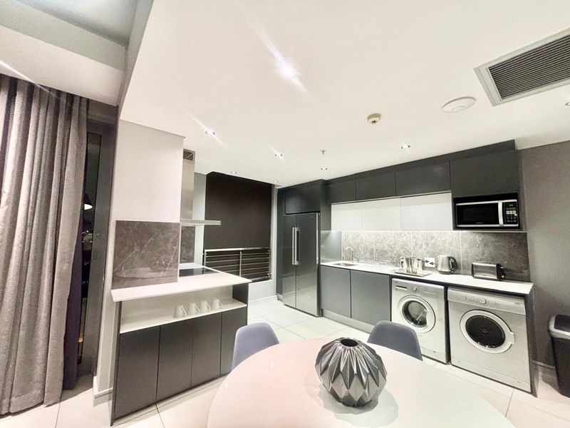 Modern open-plan kitchen featuring sleek dark cabinetry, stone backsplash, integrated appliances, and built-in laundry facilities in a well-maintained contemporary hotel suite.