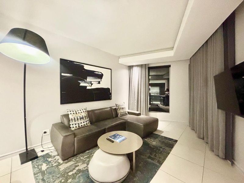 A modern hotel living room featuring tiled flooring, recessed ceiling details, and floor-to-ceiling curtains.