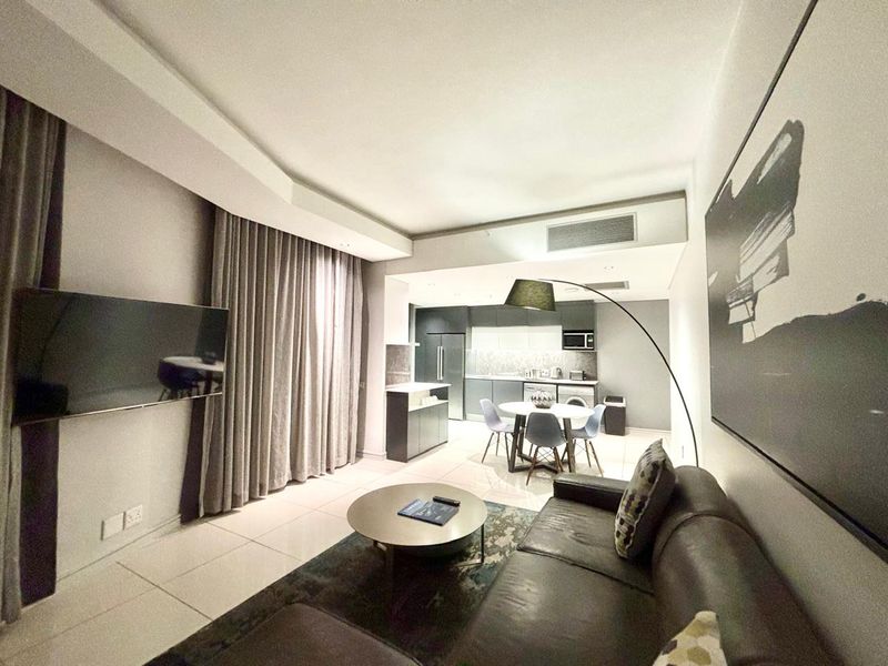 Modern open-plan hotel suite featuring a sleek kitchen with built-in cabinetry, tiled flooring, and integrated air conditioning, seamlessly flowing into a comfortable living area.