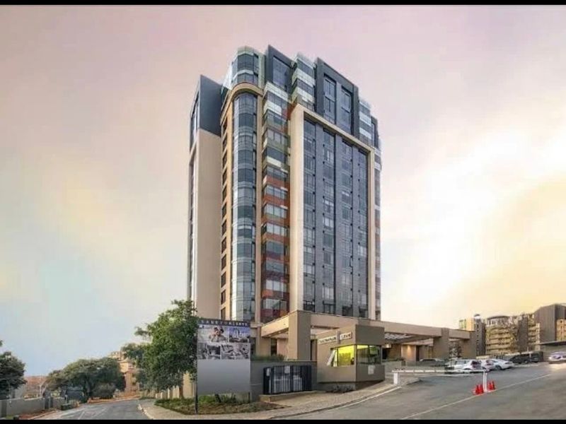 A modern multi-story apartment building featuring a glass facade, secure gated entrance, and a paved driveway approach.