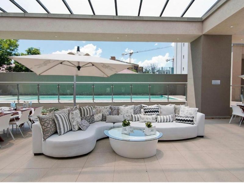 A spacious covered patio featuring tiled flooring, a glass roof canopy, and a modern glass balustrade overlooking a swimming pool.