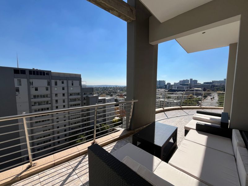 A spacious covered balcony featuring tiled flooring, a stainless steel balustrade, and expansive urban views.