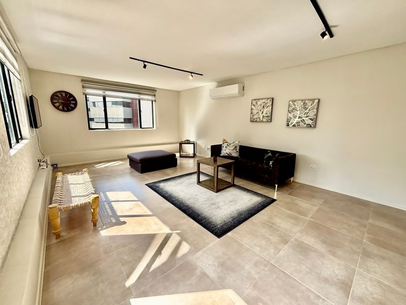 A bright and spacious living room featuring neutral tiled flooring, modern track lighting, and a wall-mounted split-system air conditioner.