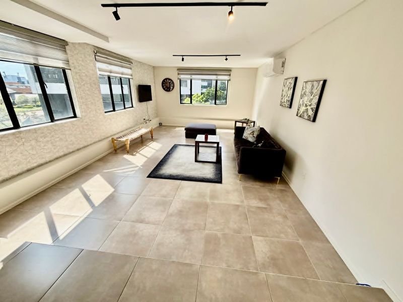 Spacious living area featuring large windows with roller blinds, neutral tiled flooring, track lighting, and a wall-mounted air conditioning unit.