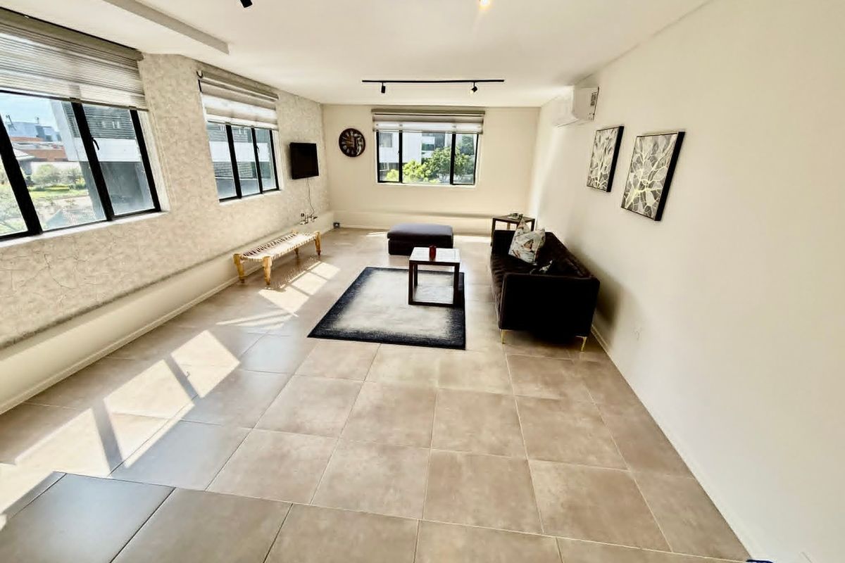Spacious living area featuring large windows with roller blinds, neutral tiled flooring, track lighting, and a wall-mounted air conditioning unit.