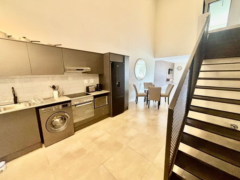 Modern open-plan kitchen and dining area featuring sleek dark cabinetry, integrated appliances, tiled flooring, and a striking industrial-style staircase.