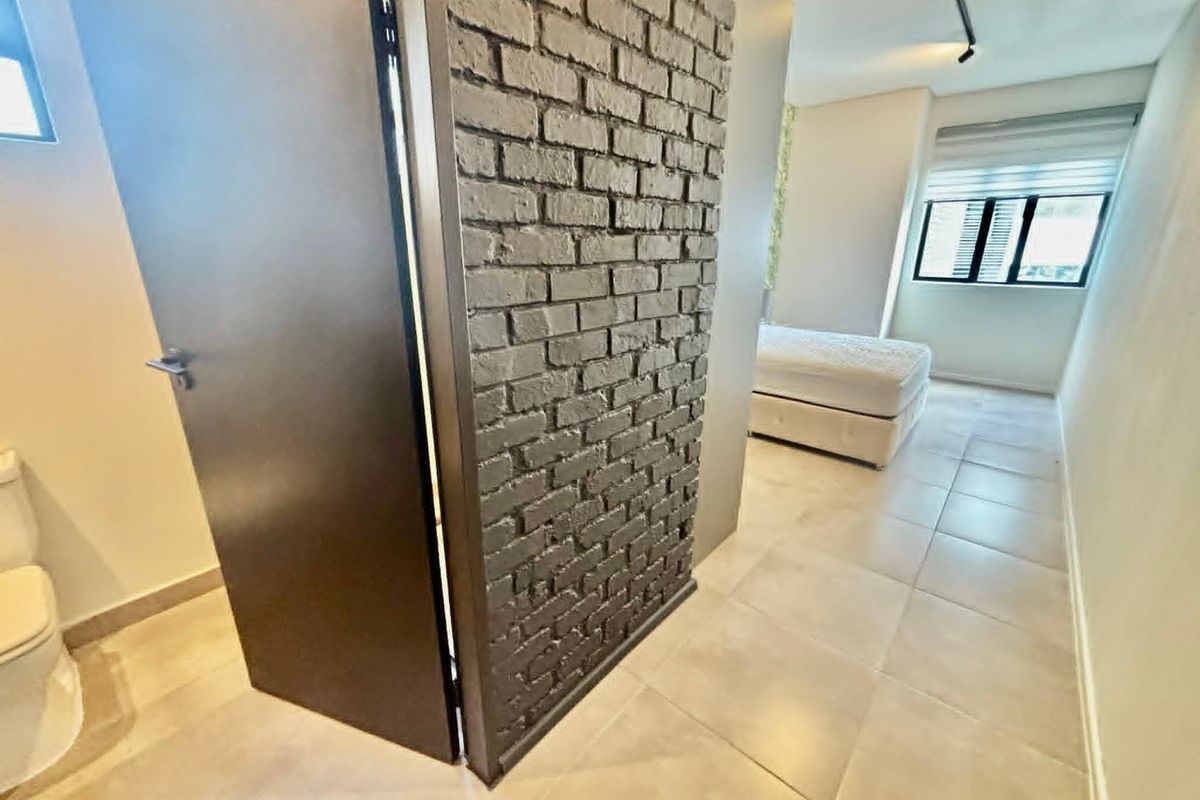 Modern bedroom featuring an exposed brick accent wall, neutral tiled flooring, and track lighting, creating a contemporary and clean living space.