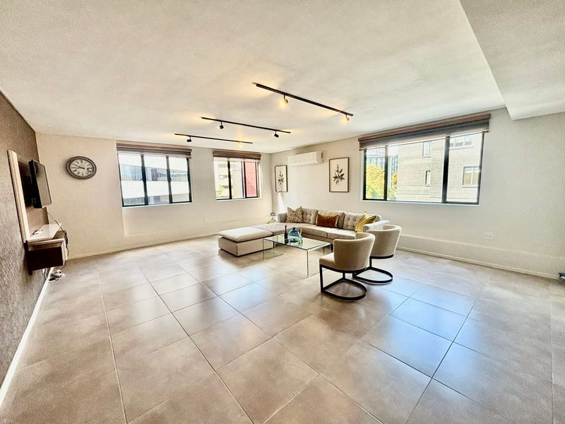 Spacious living area featuring large windows, neutral tiled flooring, modern track lighting, and a wall-mounted air conditioning unit.