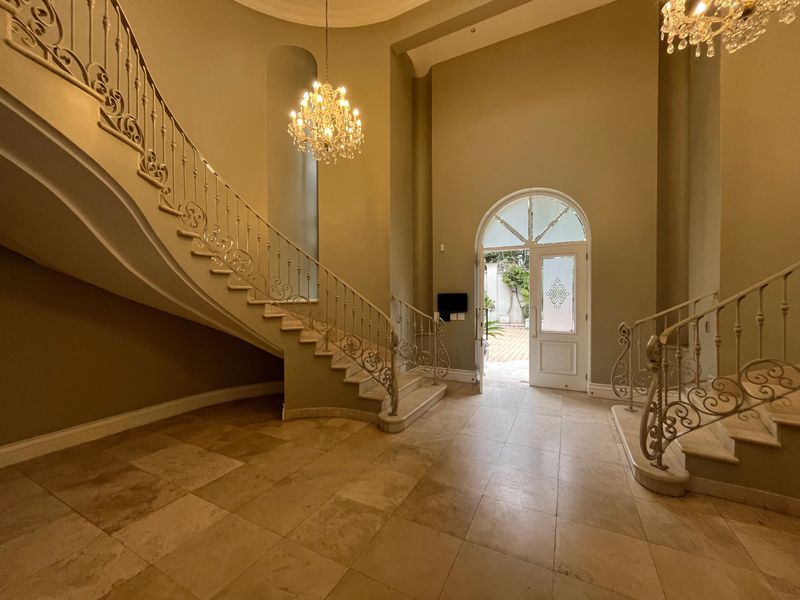 Grand double-volume entrance hall featuring elegant stone flooring, a sweeping staircase with ornate wrought iron balustrades, and dual crystal chandeliers.