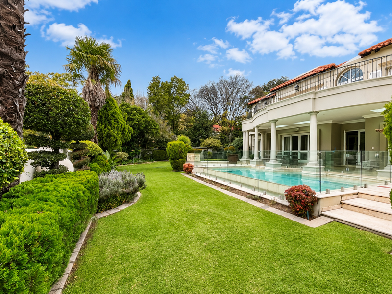A beautifully landscaped rear garden featuring a pristine swimming pool, glass balustrade, and a covered patio area with elegant stone columns.